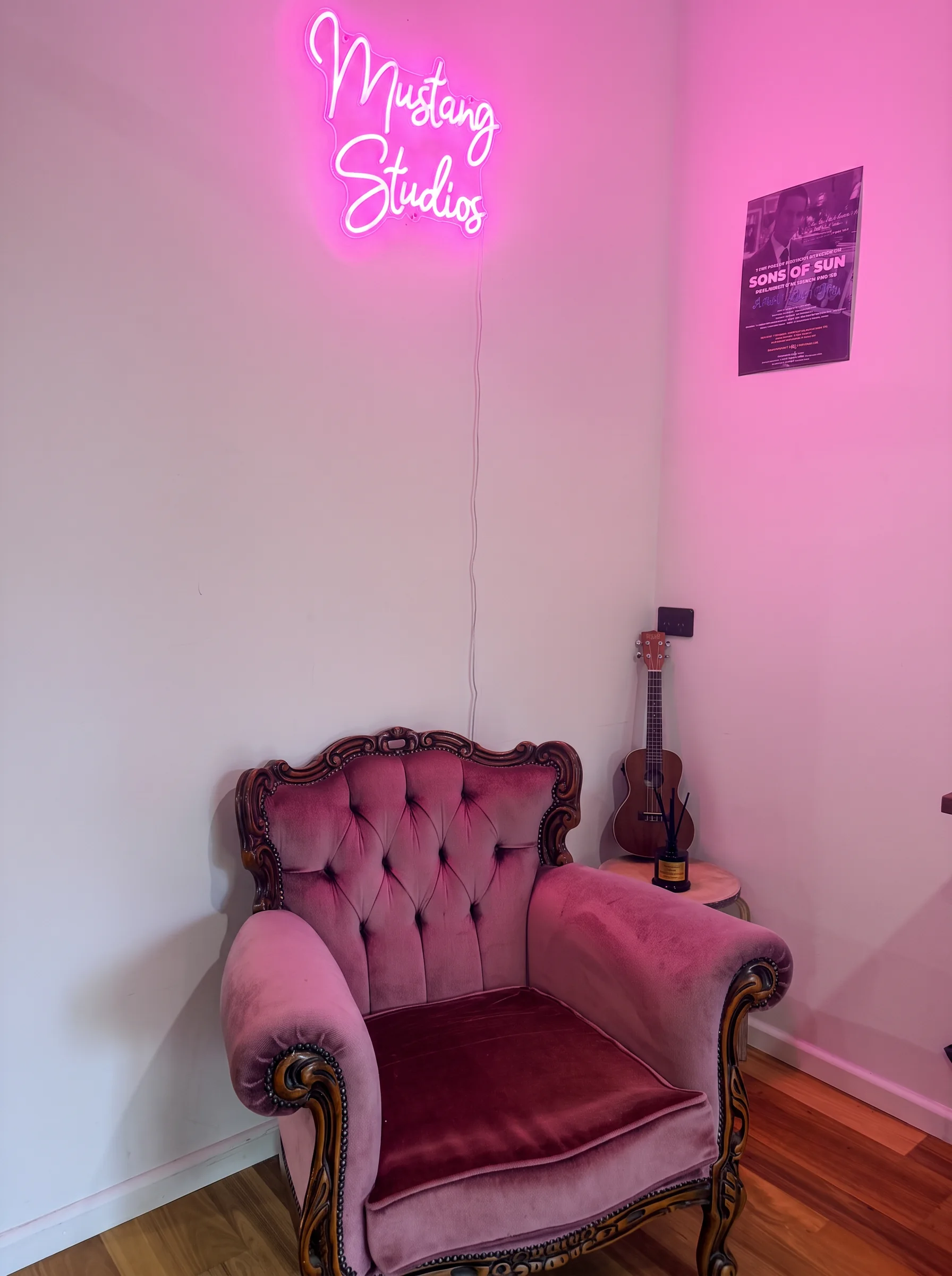 Pink neon 'Mustang Studios' sign glowing above a velvet armchair, acoustic guitar and gig poster