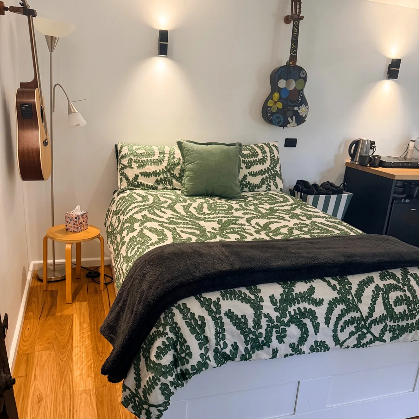 Queen bed with green patterned bedding, reading lamps and guitars hanging on the wall